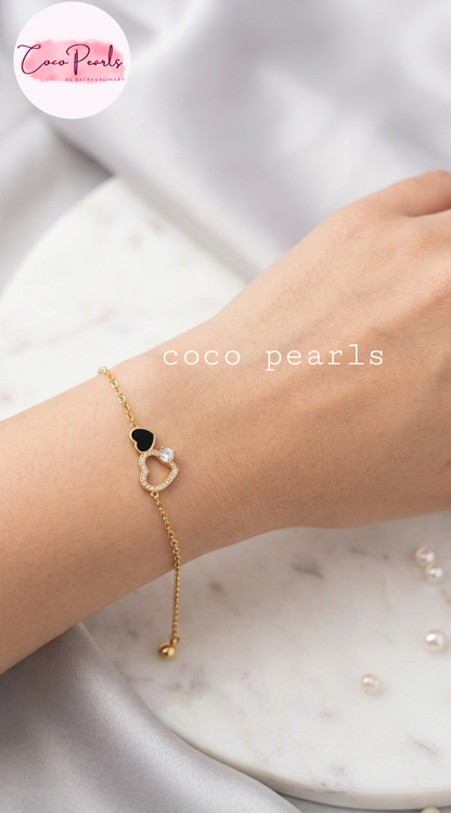 Coco pearls stainless Steel anti tarnish gold chain bracelet with a black heart and a heart-shaped pearl charm, worn on a person's wrist. The background consists of a white marble tray and soft white fabric.