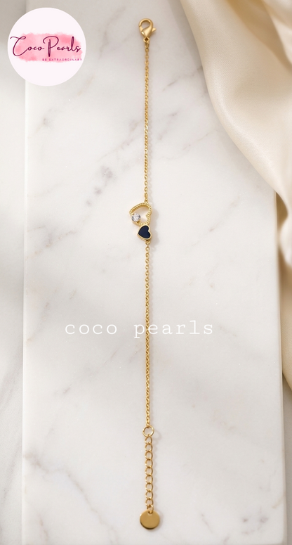 Coco pearls stainless Steel anti tarnish delicate gold chain bracelet with a small gold heart, a white pearl, and a dark blue heart charm, displayed on a white marble slab with a cream satin cloth in the corner.