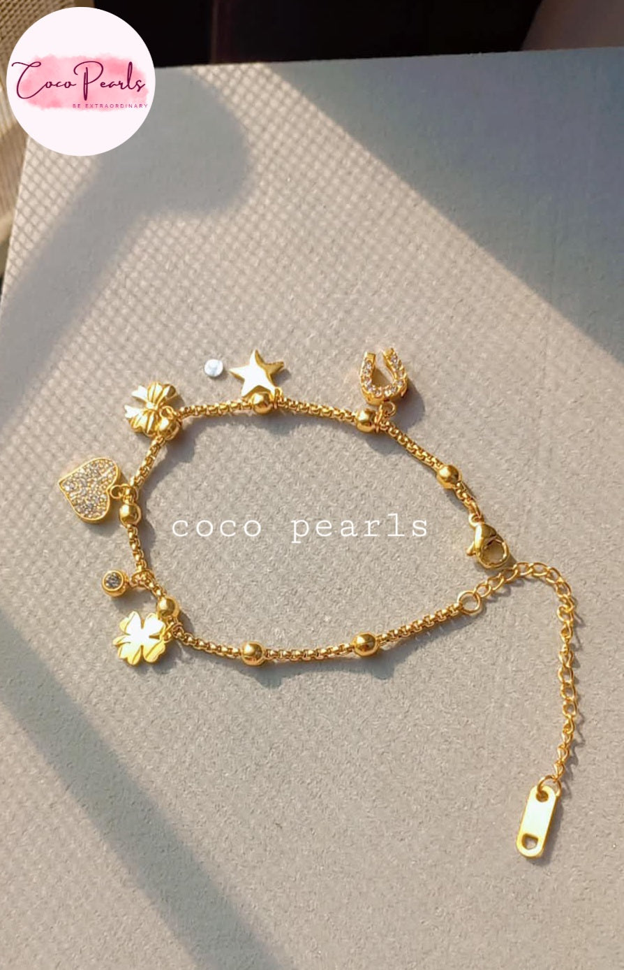 Coco pearls stainless Steel anti tarnish delicate gold charm bracelet with floral, star, and heart charms, displayed on a textured beige surface with soft sunlight shadows. 