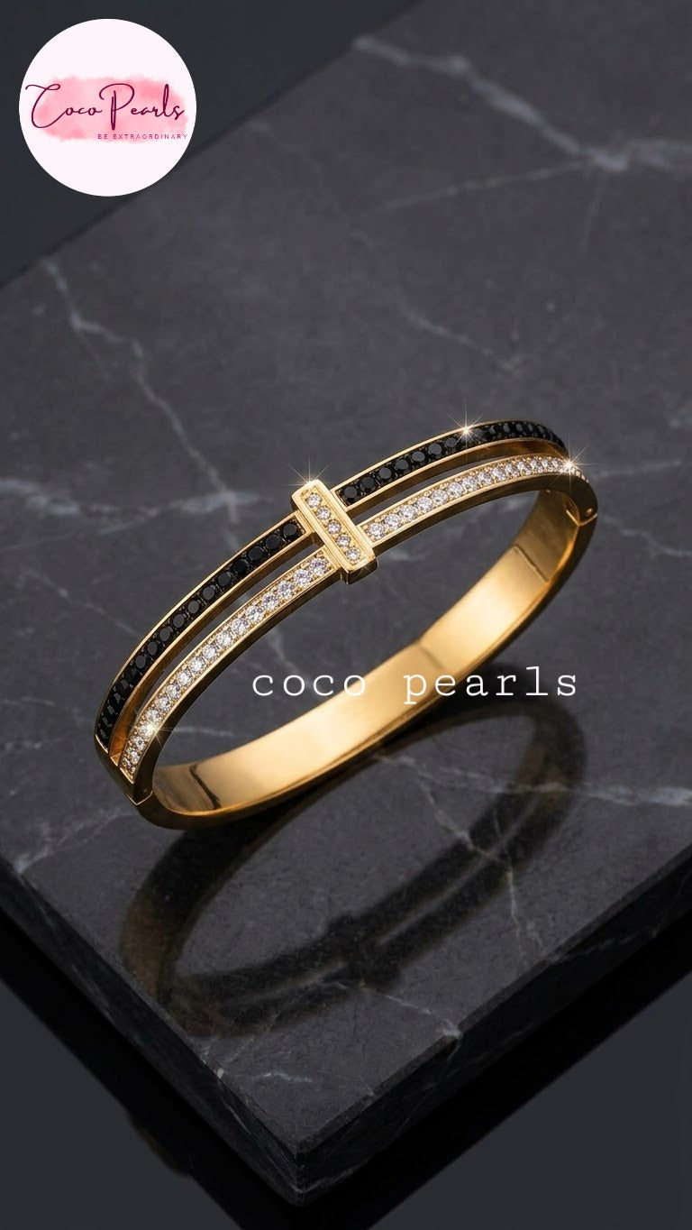 Coco pearls stainless Steel anti tarnish gold-plated bangle bracelet with black enamel sections and rows of clear pavé-set stones, displayed on a dark marble slab.