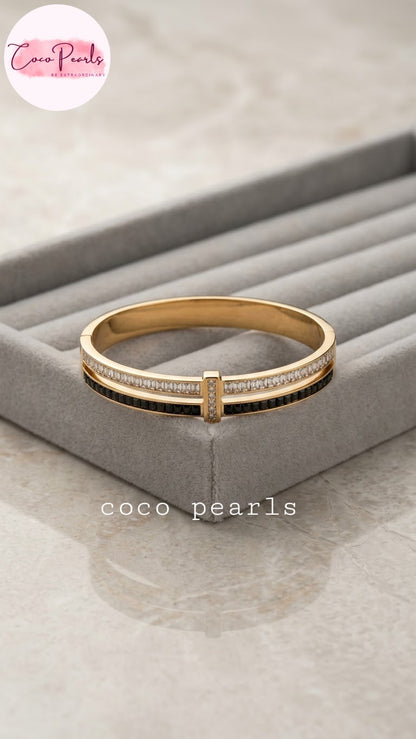 Coco pearls stainless Steel anti tarnish gold-plated bangle bracelet with a central black stripe and rows of clear stones on the front, displayed on a gray jewelry organizer tray.
