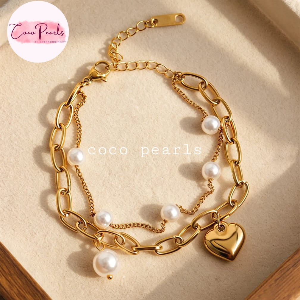 Coco pearls stainless Steel anti tarnish gold-tone layered chain bracelet with white pearl accents and a polished heart charm, displayed on a beige textured surface