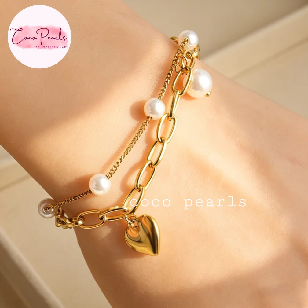 Coco pearls stainless Steel anti tarnish close-up of a gold chain bracelet with white freshwater pearls and a dangling heart charm, shown worn on a person's wrist.