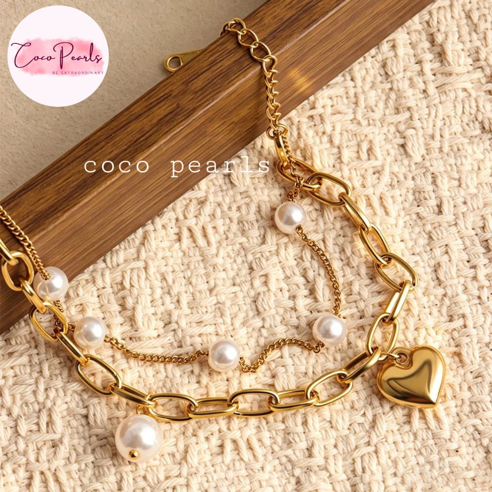 Coco pearls stainless Steel anti tarnish gold-tone layered chain bracelet with white faux pearls and a polished heart charm, displayed on a textured cream fabric
