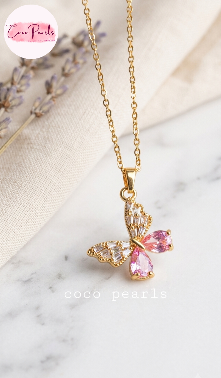 Coco pearls stainless Steel anti tarnish Gold butterfly pendant necklace with pink teardrop crystals and clear rhinestones, displayed on a marble surface with a cream fabric backdrop.