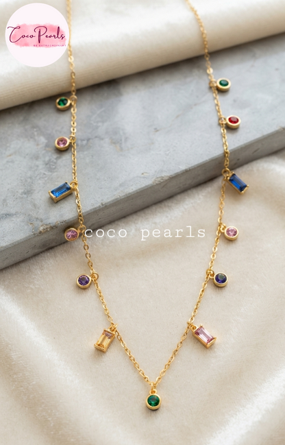 Coco pearls stainless Steel anti tarnish gold chain necklace with multiple colorful gemstone charms, displayed on a beige satin fabric with a gray stone slab prop. The necklace features round, rectangular, and oval gemstones in green, blue, pink, purple, and red.