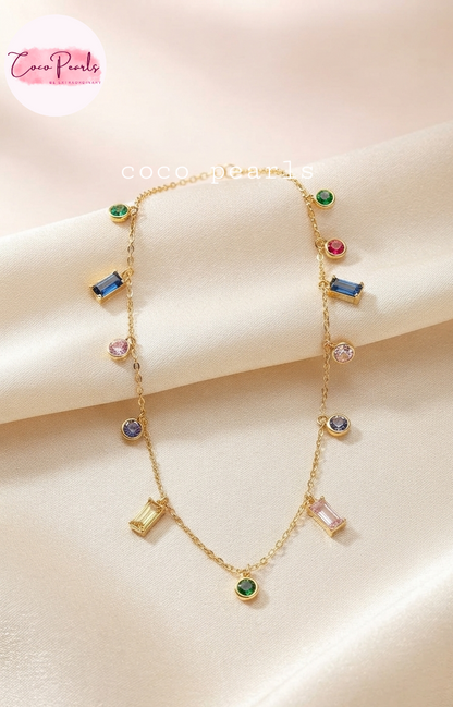 Coco pearls stainless Steel anti tarnish gold chain necklace with multiple colorful gemstone charms, displayed on a soft beige satin fabric. The necklace features various gemstone shapes including round, oval, and rectangular bezels in green, blue, pink, and clear stones.