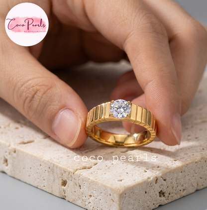 Coco pearls stainless Steel anti tarnish gold ring with a round clear gemstone held between fingers over a textured beige stone slab.