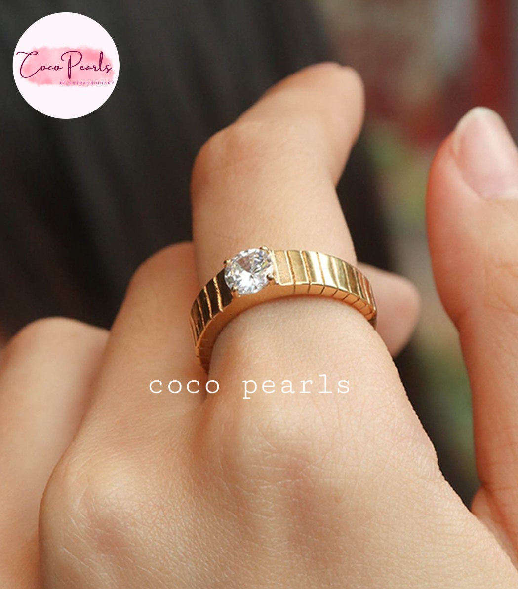 Coco pearls stainless Steel anti tarnish close-up of a hand wearing a gold ring with a single clear round gemstone. The ring has a textured, segmented band.