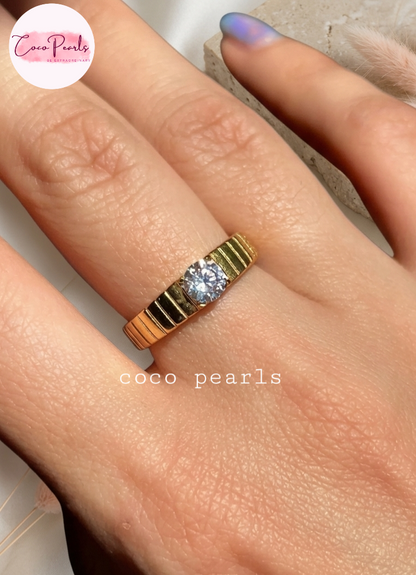 Coco pearls stainless Steel anti tarnish close-up of a hand wearing a gold ring with a central clear gemstone. The ring has a textured, ribbed gold band.