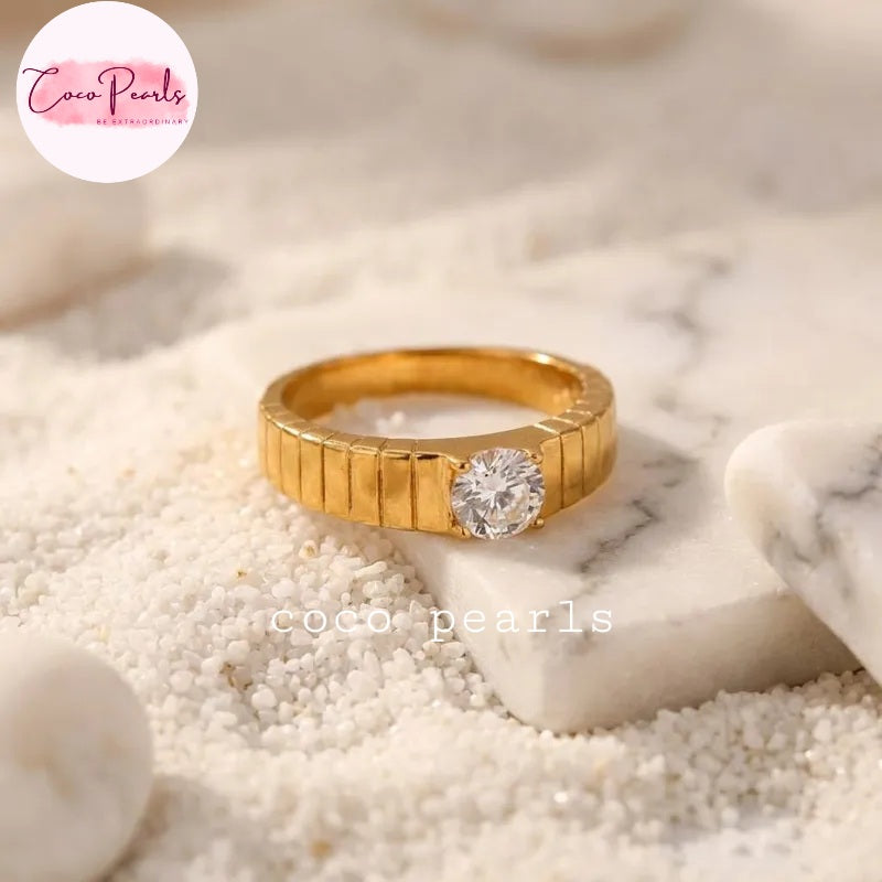 Coco pearls stainless Steel anti tarnish gold ring with a textured, ribbed band and a round clear gemstone, resting on white sand and a small white marble slab.