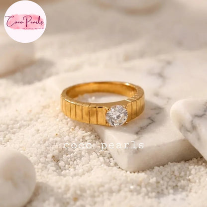 Coco pearls stainless Steel anti tarnish gold ring with a textured, ribbed band and a round clear gemstone, resting on white sand and a small white marble slab.