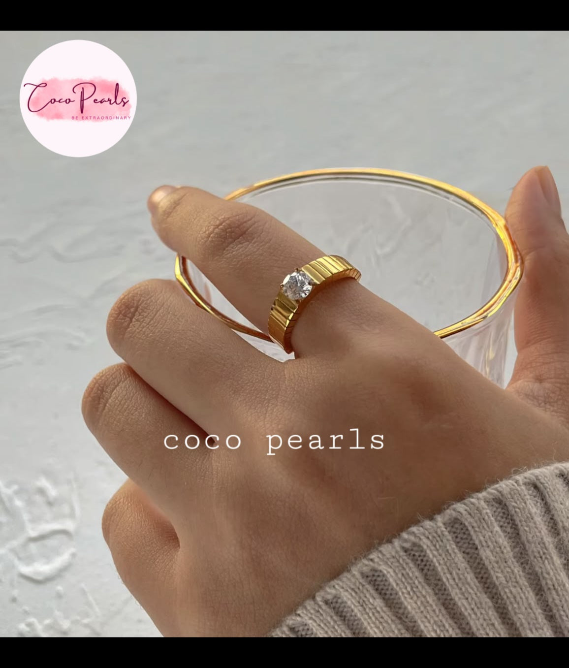 Coco pearls stainless Steel anti tarnish close-up of a hand wearing a gold ring with a clear central gemstone, holding a clear glass.