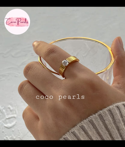 Coco pearls stainless Steel anti tarnish close-up of a hand wearing a gold ring with a clear central gemstone, holding a clear glass.