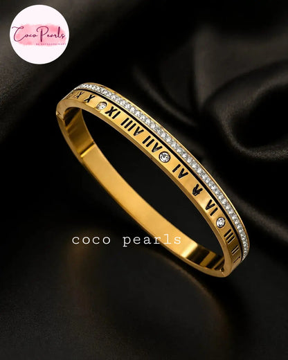 Coco pearls stainless Steel anti tarnish gold bangle bracelet with Roman numerals and clear rhinestones, displayed on a black satin background. 