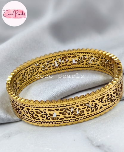 Coco pearls stainless Steel anti tarnish gold-plated filigree bangle bracelet with intricate cut-out patterns, displayed on a soft white fabric background.