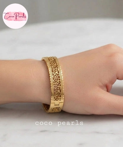 Coco pearls stainless Steel anti tarnish gold-toned filigree bangle bracelet with a geometric clasp, shown worn on a person's wrist against a light marble background.