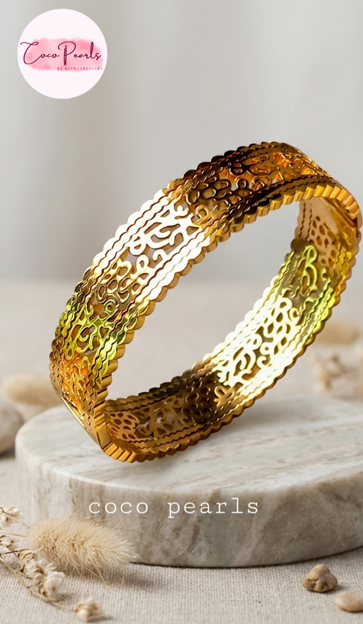 Coco pearls stainless Steel anti tarnish wide, gold-toned bangle bracelet with intricate cutout filigree patterns, displayed on a round marble pedestal.