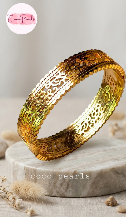 Coco pearls stainless Steel anti tarnish wide, gold-toned bangle bracelet with intricate cutout filigree patterns, displayed on a round marble pedestal.
