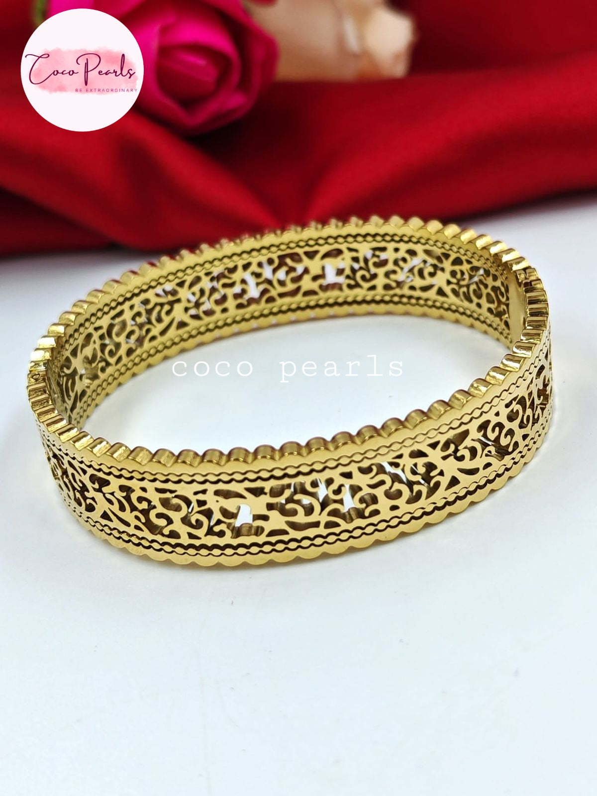Coco pearls stainless Steel anti tarnish gold-plated filigree bangle bracelet with intricate cut-out patterns, displayed on a white surface with a red satin fabric and a pink rose in the background.