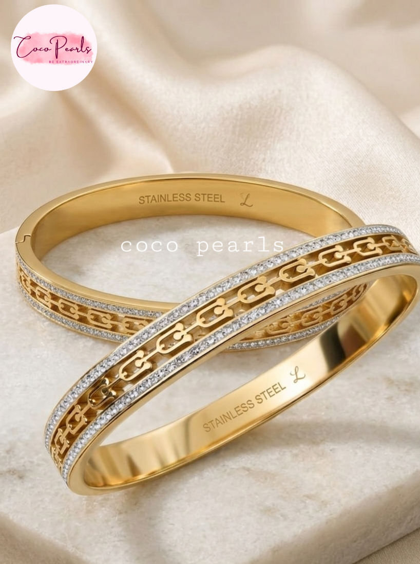 Coco pearls stainless Steel anti tarnish Two stainless steel bangle bracelets with interlocking chain motifs, one adorned with clear pavé stones, displayed on a cream satin fabric background.