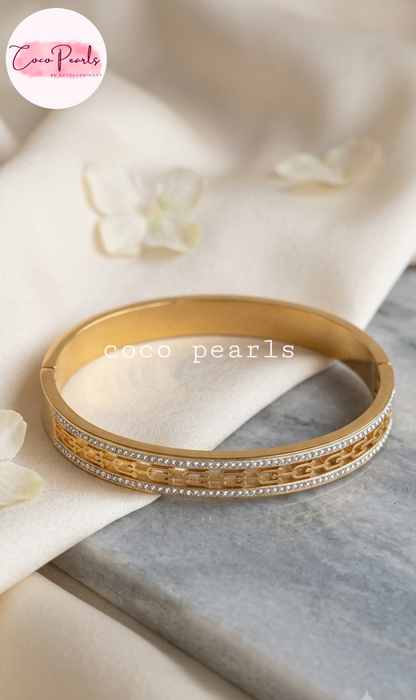 Coco pearls stainless Steel anti tarnish gold bangle bracelet with intricate chain link detailing and rows of small clear stones, displayed on a cream fabric