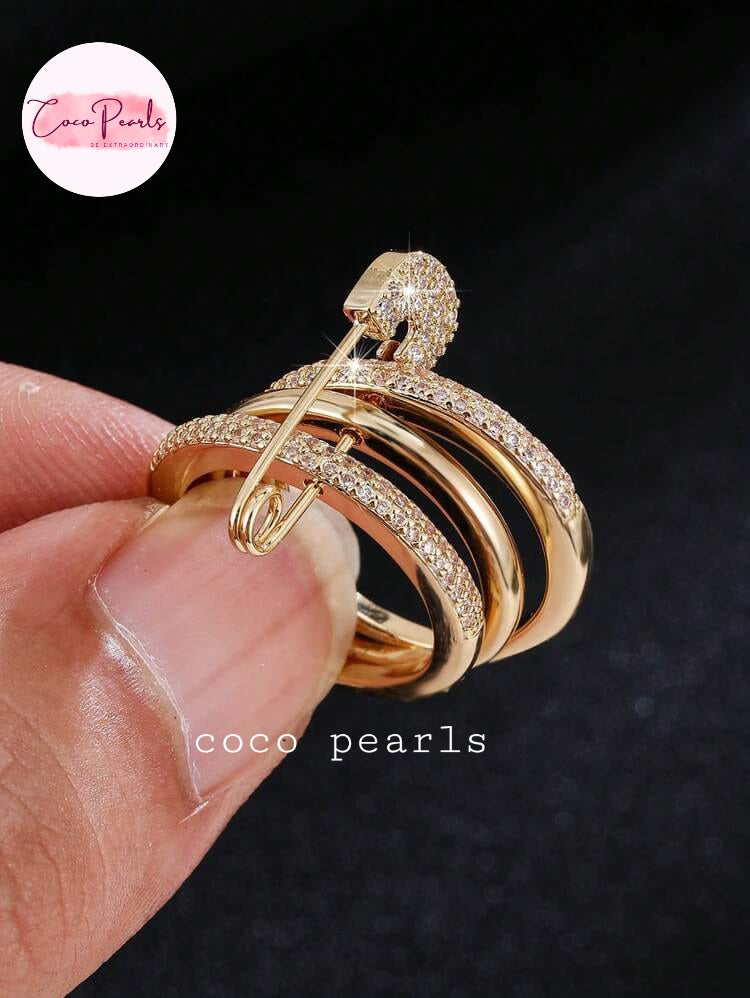 Coco pearls stainless Steel anti tarnish close-up of a hand holding a gold safety-pin style ring with multiple curved bands, some set with small clear stones, against a dark background. 