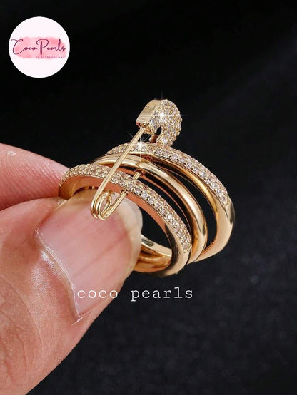 Coco pearls stainless Steel anti tarnish close-up of a hand holding a gold safety-pin style ring with multiple curved bands, some set with small clear stones, against a dark background. 