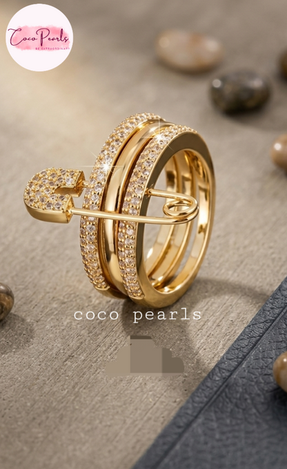 Coco pearls stainless Steel anti tarnish gold safety-pin style ring with multiple bands, some encrusted with clear stones, displayed on a textured neutral surface with decorative stones.