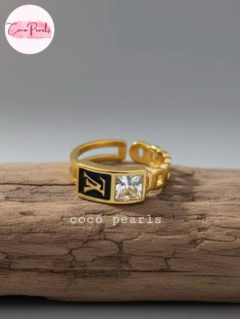 Coco pearls stainless Steel anti tarnish gold-tone open-shank ring with a rectangular front face featuring a black inlay with a gold LV monogram and a clear square gemstone, displayed on a piece of weathered wood 