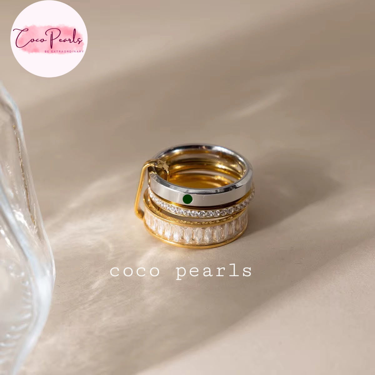 Coco pearls stainless Steel anti tarnish stack of three rings displayed on a beige fabric surface. The rings feature a gold and silver finish, with one ring adorned with a small green stone and another with clear crystal stones
