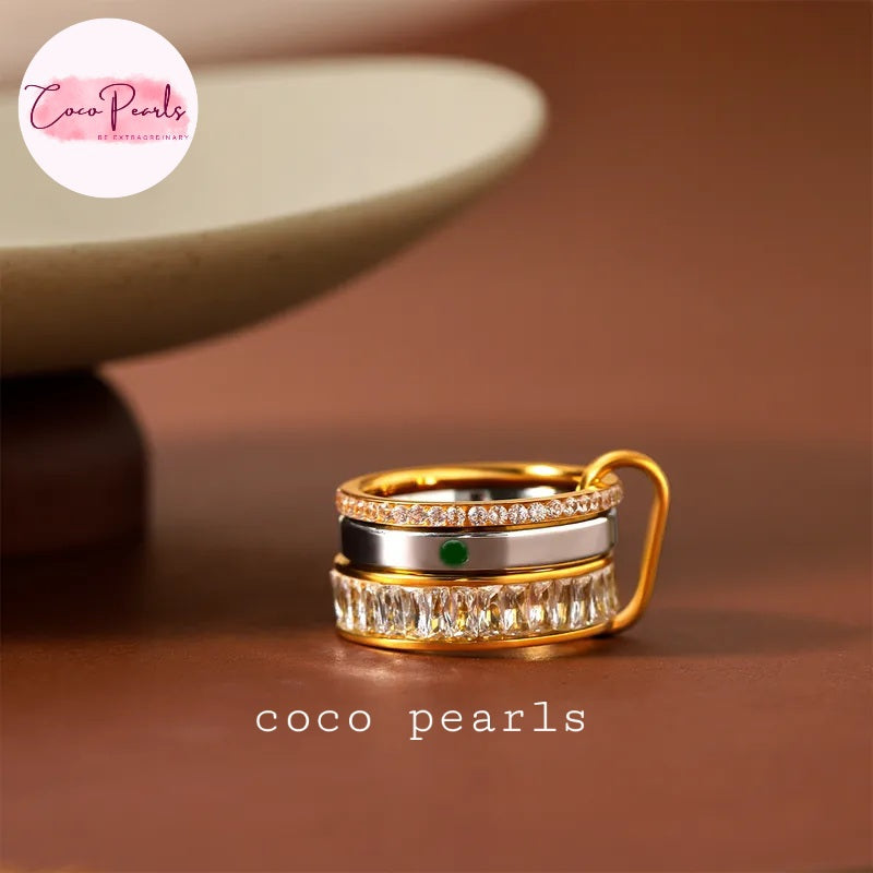 Coco pearls stainless Steel anti tarnish gold-plated ring with stacked bands, featuring a top band of small clear stones, a middle band with a single green gemstone, and a bottom band of larger rectangular clear stones, shown on a brown surface
