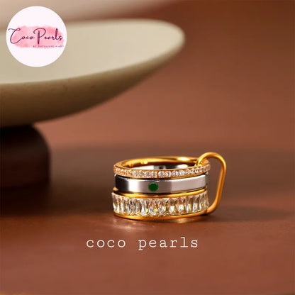 Coco pearls stainless Steel anti tarnish gold-plated ring with stacked bands, featuring a top band of small clear stones, a middle band with a single green gemstone, and a bottom band of larger rectangular clear stones, shown on a brown surface