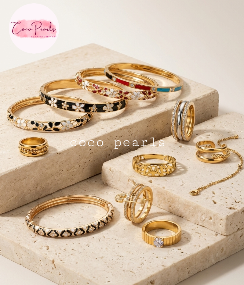 A collection of Coco Pearls gold bangles and rings displayed on beige stone blocks against a cream background. The jewelry features gold-plated metals with black floral enamel, white floral enamel, red enamel, turquoise enamel, and clear crystal details.
