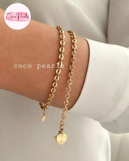 Coco pearls stainless Steel anti tarnish close-up of a person's wrist wearing two delicate gold bracelets, one with a curb link style and one with a floral pattern and a dangling sunburst heart charm.