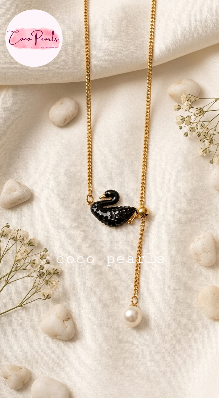 Coco pearls stainless Steel anti tarnish gold-tone dangle necklace with a black swan pendant and a dangling pearl, styled on a cream fabric background