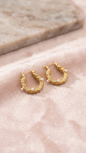 Coco pearls stainless Steel anti tarnish pair of textured gold hoop earrings adorned with small white pearls, displayed on a soft pink fabric surface with a blurred beige stone background.