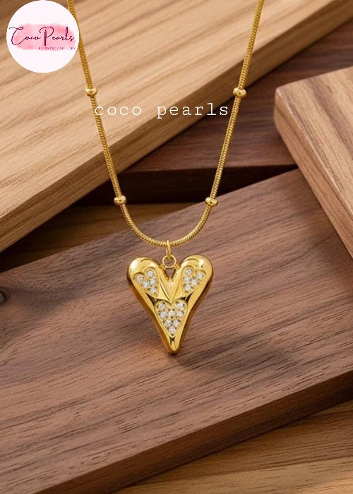 Coco pearls stainless Steel anti tarnish gold heart-shaped pendant necklace with clear rhinestones on a gold chain, displayed on a wooden background.