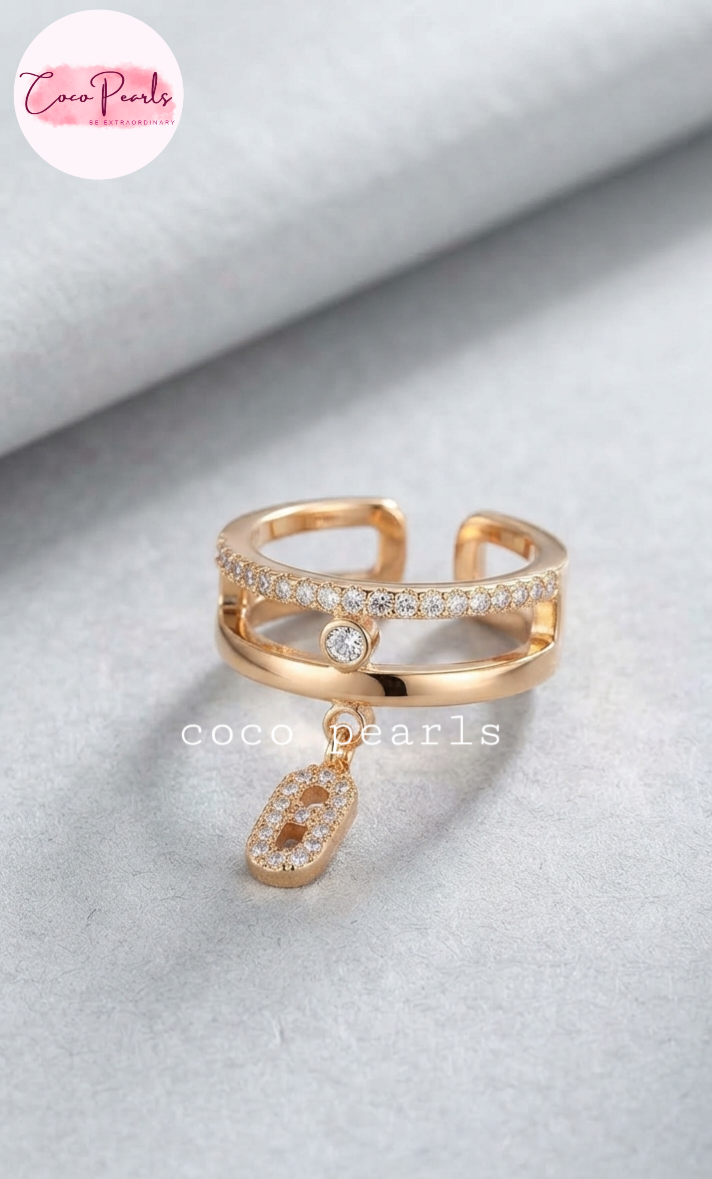 Coco pearls stainless Steel anti tarnish gold adjustable ring with a double-band design, one band paved with clear stones and a central round stone, and a dangling oval stone charm, displayed on a textured light gray surface.