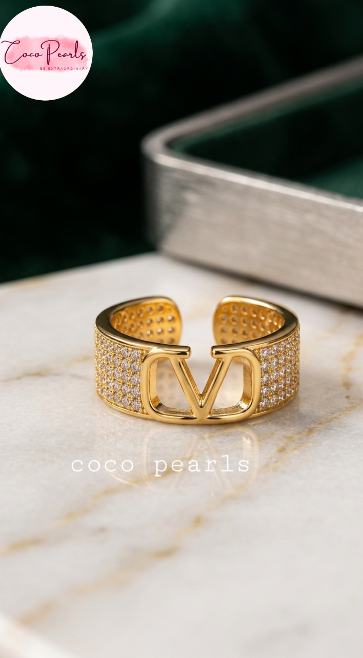 Coco pearls stainless Steel anti tarnish gold-toned cuff ring with pavé-set clear stones and a large interlocking 'V' motif on the front, displayed on a light marble surface with a dark green velvet jewelry box