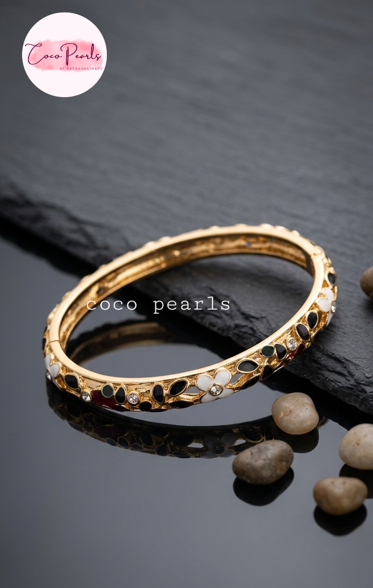Coco pearls gold-plated bangle bracelet with black and white floral inlays, displayed on a dark slate surface with small decorative stones. 