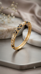  Coco pearls  gold plated bangle bracelet with black and white floral engravings, displayed on a gray tray with a beige fabric background.