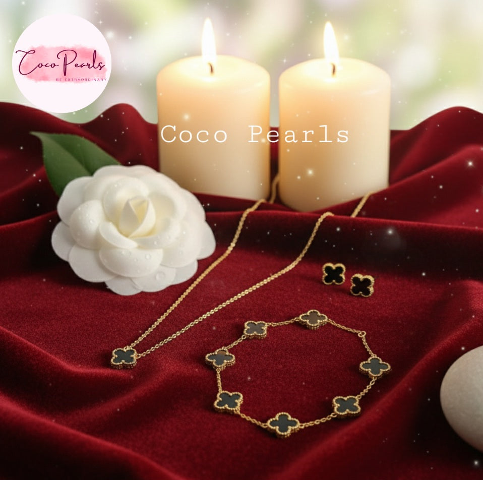Coco Pearls Stainless Steel Anti Tarnish Gold black clover dotted jewelry set with necklace and earrings on a red fabric background with candles and a flower.