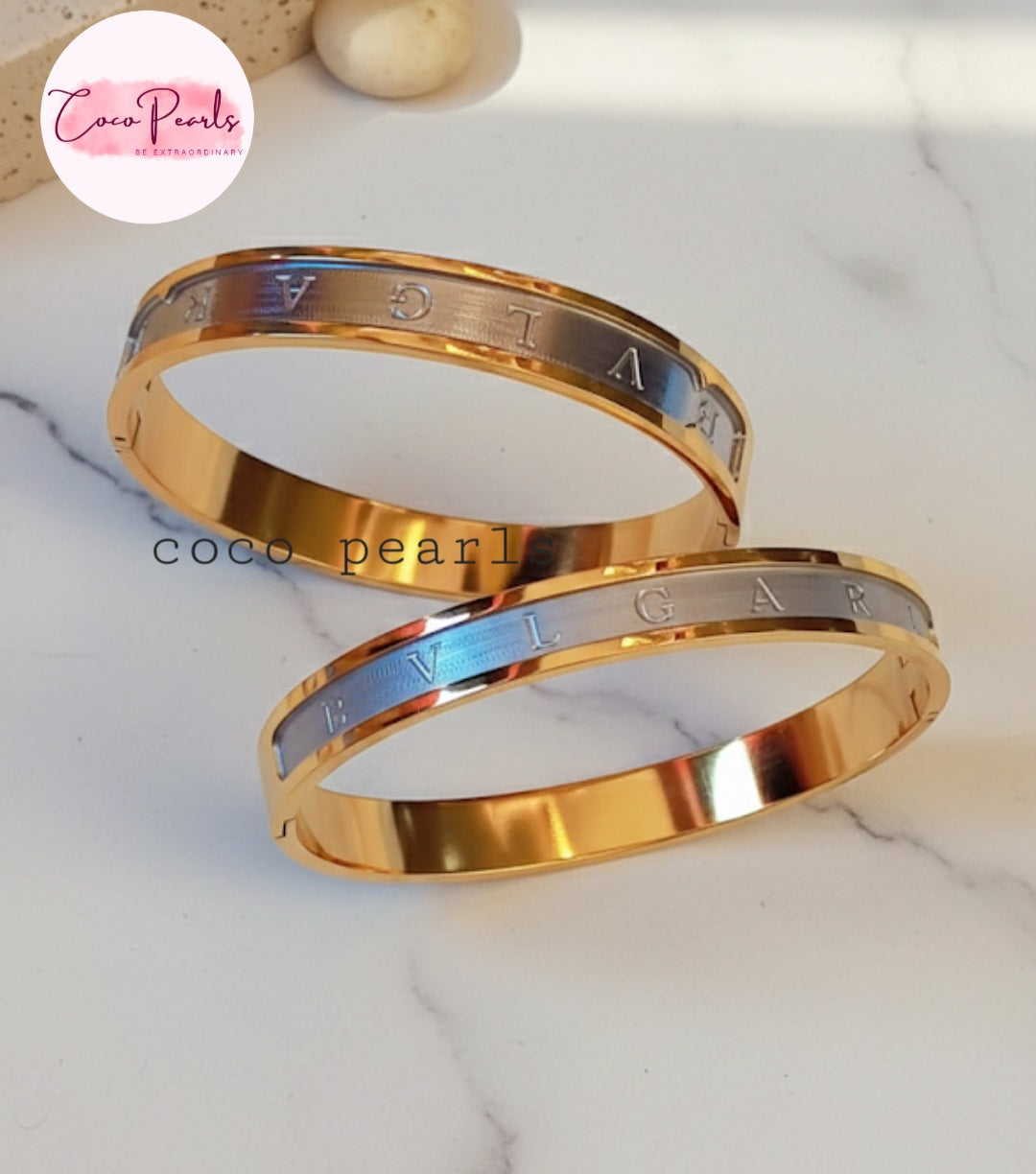 Coco Pearls Stainless Steel Anti Tarnish Two gold and silver coco glitz handcuff bracelets with 'Bulgari' engraving on a white marble surface