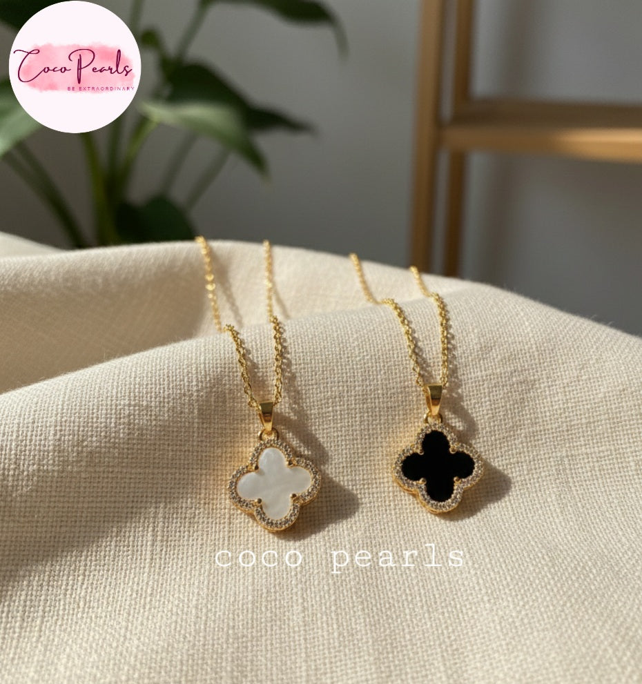 Coco Pearls Stainless Steel Anti Tarnish Two sided diamond studded gold necklaces with clover-shaped pendants one side black one side white on a beige surface.