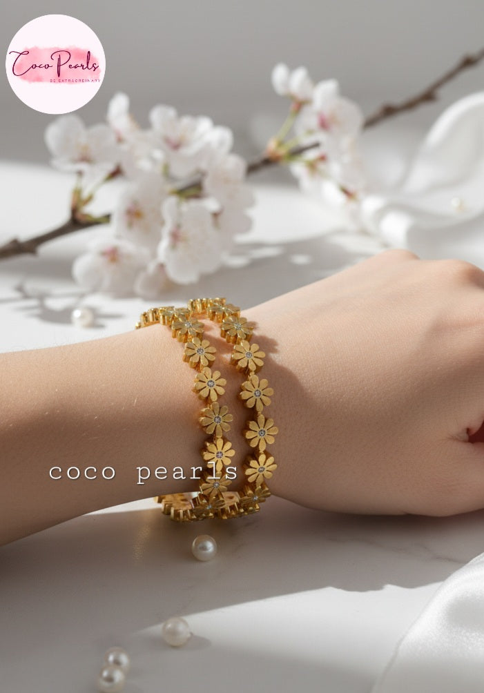 Coco Pearls Stainless Steel Anti Tarnish Gold daisy bloom floral bangle bracelet with floral design on a wrist, cherry blossoms in the background