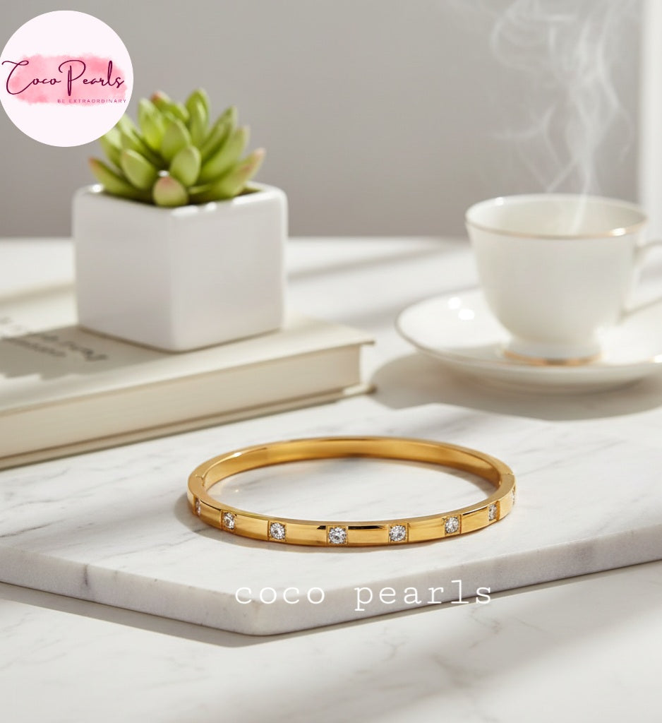 Coco Pearls Stainless Steel Anti tarnish Gold diamond radiance handcuff bangle bracelet with crystals displayed on a marble surface with a cup of tea and plant in the background
