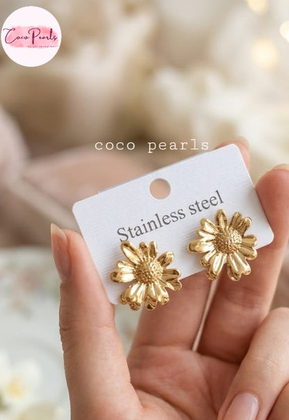  coco pearls stainless steel jewelry pakistan anti tarnish waterproof Golden daisy flower-shaped earrings held by a hand with a blurred background.