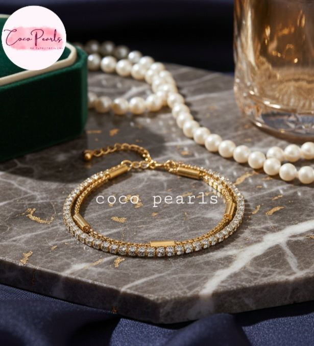 Coco Pearls Stainless steel Anti Tarnish Golden stream link bracelet with double layer chain. a diamond studded line and golden chain displayed on a marble surface with pearls and a glass.