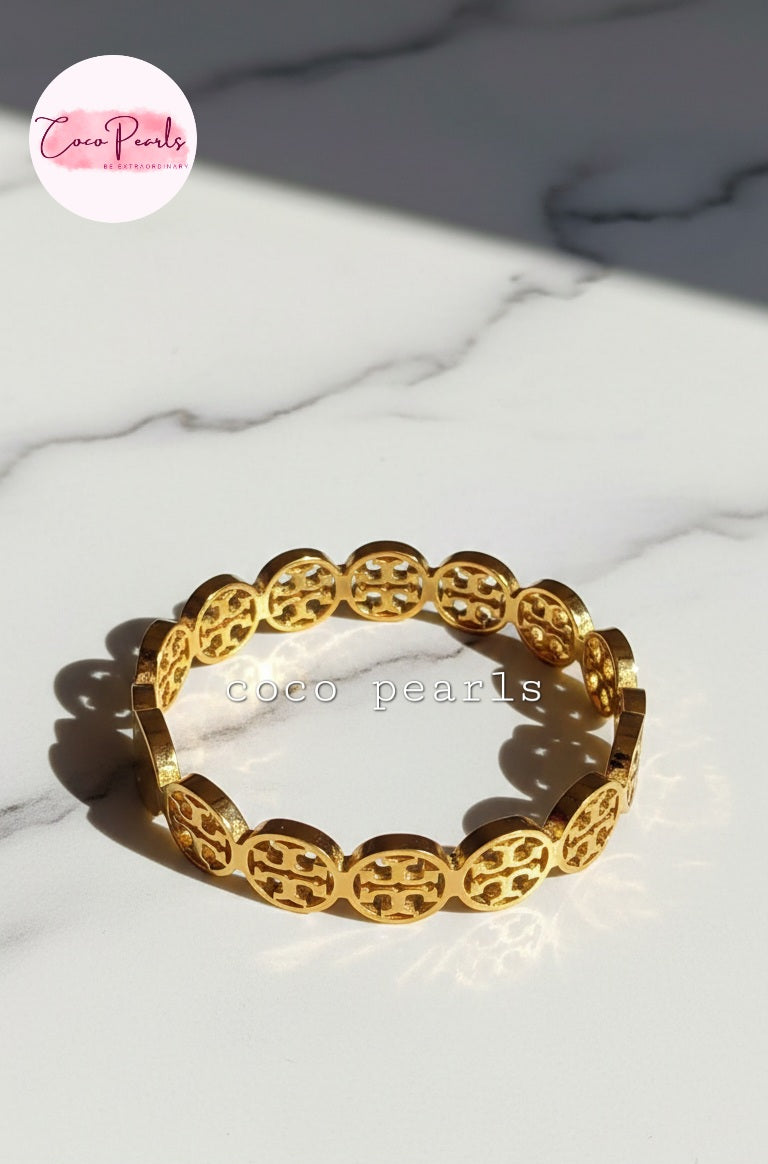 coco pearls stainless steel anti tarnish gold interlocking logo handcuff with all over logos connected displayed on a marble surface